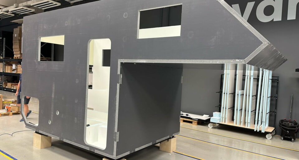 Ein großer, unfertiger Pickup-Camper mit markant-eckigem Design – ähnlich dem Pickupboxx Allround-Talent – steht drinnen auf Holzklötzen. Sichtbare Fenster und eine offenstehende Türöffnung lassen grenzenlose Freiheit erahnen. Im Hintergrund liegen verstreute Werkzeuge und Ausrüstungsgegenstände, die echtes Offroad-Feeling vermitteln. Dieser individuelle Custom Camper ist die ideale Basis für alle, die ihren Traum vom eigenen Offroad-Camper oder Pickup Camper verwirklichen möchten. Travelboxx - Pickup Wohnkabinen