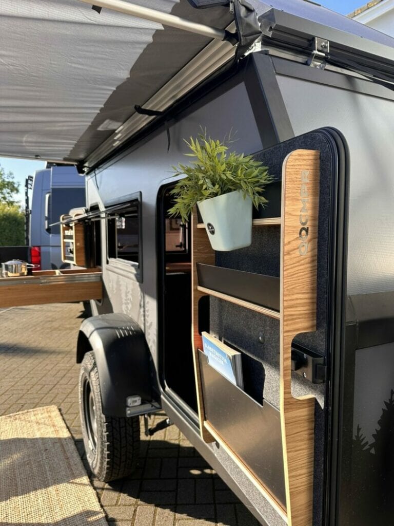 Nahaufnahme eines schwarzen Teardrop-Campers mit geöffneter Seitentür – ideal als Pickup Camper oder kompakter Offroad Camper. Im Innenraum bieten Regale Platz für Pflanzen und Zeitschriften, während am Heck die Outdoor-Küche ausgezogen ist. Ein ausklappbares Sonnensegel spendet Schutz vor Sonne und Regen. Dieser maßgefertigte Custom Camper überzeugt als vielseitiges Allround-Talent und ermöglicht grenzenlose Freiheit auf jedem Abenteuer, egal ob Offroad-Trip oder entspannter Campingurlaub mit dem Pickup Truck. Travelboxx - Pickup Wohnkabinen
