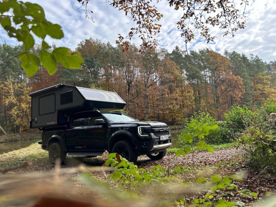 Ein schwarzer Pickupboxx-Truck mit einem Custom-Camper im Ladebett steht auf einem Offroad-Pfad neben einem Teich, umgeben von herbstlichen Bäumen und viel Grün. Dieses Offroad-Camper-Setup verkörpert das echte Allround-Talent für grenzenlose Freiheit – perfekt für Abenteuerlustige, die einen Pickup Camper, der sowohl in der Wildnis als auch abseits befestigter Straßen zu Hause suchen ist. Das Ganze wird unter einem teils bewölkten Himmel eindrucksvoll in Szene gesetzt. Travelboxx - Pickup Wohnkabinen