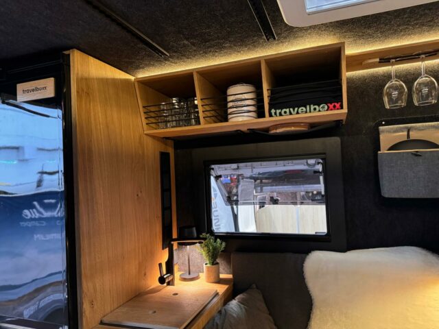 Gemütliches Pickup Camper-Interieur mit einem kleinen Schreibtisch aus Holz, Lampe, Pflanzen, Ablagen mit Geschirr und einer „Allround-Talent“ Travelboxx. Über dem eleganten Custom-Camper-Innenraum hängen Weingläser, während durch das Fenster grenzenlose Freiheit und Offroad-Abenteuer in der Natur sichtbar werden – ideal für individuelle Camper-Träume und anspruchsvolle Reisende auf der Suche nach dem perfekten Offroad-Camper.​. Travelboxx - Pickup Wohnkabinen