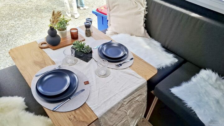 Ein gemütlich gedeckter Esstisch für zwei Personen mit dunkelblauen Tellern, Gläsern, Besteck, einem beigen Tischläufer, Topfpflanzen, Kerzen und einer modernen Vase - die perfekte Ausstattung für deinen Pickup Camper oder Offroad Camper. Dunkle Kissen und weiße Kunstfelldecken sorgen dabei für zusätzlichen Komfort und ein stilvolles Ambiente in deinem Custom Camper. Ideal für unvergessliche Abenteuer mit dem Wohnanhänger oder der Travelboxx Quest!. Travelboxx - Pickup Wohnkabinen