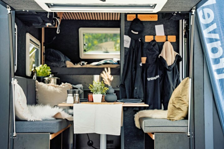 Innenraum eines gemütlichen Pickup Campers: Der maßgefertigte Offroad Camper überzeugt mit einem einladenden Essbereich, bestehend aus einem stabilen Tisch und gepolsterten Sitzbänken. Grüne Pflanzen und weiche Kissen sorgen für eine wohnliche Atmosphäre. An den Wandhaken hängen Jacken und Hüte griffbereit. Im hinteren Teil des Custom Campers befindet sich ein komfortables Bett direkt am Fenster, das einen tollen Ausblick ermöglicht. Durch die großen Fensterflächen wird der Innenraum des Travelboxx Quest von natürlichem Licht durchflutet - perfekt für alle, die auf ihren Outdoor-Abenteuern Wert auf Komfort und Gemütlichkeit legen. Travelboxx - Pickup Wohnkabinen