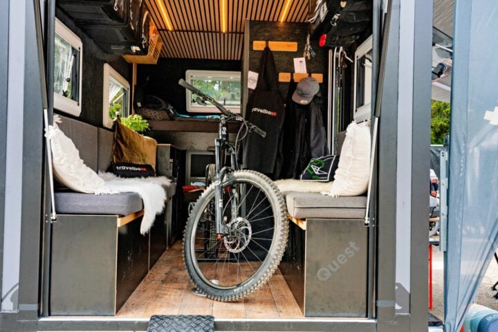 Ein Mountainbike steht im Inneren eines Travelboxx Pickup Campers, der mit gepolsterten Sitzbänken auf beiden Seiten, Kissen und an den Wänden aufgehängtem Outdoor-Equipment ausgestattet ist. Dieser maßgefertigte Offroad Camper bietet eine gemütliche und praktische Umgebung für Abenteuer in der Natur und überzeugt durch durchdachte Stauraumlösungen und robustes Design - ideal für alle, die nach einem Custom Camper oder Pickup Camper suchen. Travelboxx - Pickup Wohnkabinen