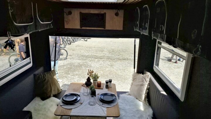 Innenraum eines Pickup Campers oder Offroad Campers mit einer gemütlichen Sitzecke: Ein stabiler Holztisch ist stilvoll mit Tellern und Gläsern gedeckt, umgeben von bequemen, gepolsterten Sitzbänken. Große Fenster bieten einen tollen Ausblick nach draußen, wo Fahrräder und Menschen zu sehen sind - perfekt für alle, die ein individuelles Camper-Erlebnis suchen. Mit diesem Custom Camper wird dein nächstes Travelboxx Quest Abenteuer unvergesslich!. Travelboxx - Pickup Wohnkabinen