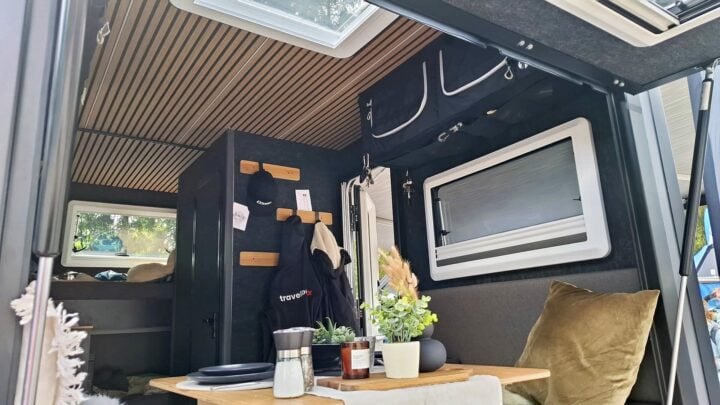 Innenraum eines modernen Pickup Campers oder Offroad Campers: Ausgestattet mit einer stilvollen Holzlamellen-Decke, einer kompakten Küchenzeile, einem Tisch mit Pflanzen und Gewürzen sowie praktischen Staufächern. Große Fenster sorgen für viel Tageslicht, während im hinteren Bereich ein gemütliches Bett zum Entspannen einlädt. An der Tür befinden sich Haken für Jacken - ideal für dein nächstes Abenteuer mit einem individuell ausgestatteten Custom Camper wie dem Travelboxx Quest. Travelboxx - Pickup Wohnkabinen