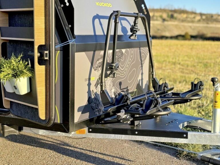 Nahaufnahme eines kompakten Pickup Campers mit modernem, aerodynamischem Design und montiertem Fahrradträger an der Front – ein echtes Allround-Talent für Abenteuerlustige. Direkt am Eingang stehen Topfpflanzen, im Hintergrund erstrecken sich weite, grüne Wiesen – perfekt für grenzenlose Freiheit auf jedem Offroad-Trip. Dieser individuelle Custom Camper vereint Komfort und Funktionalität und lässt sich ideal an Ihre Outdoor-Bedürfnisse anpassen. Travelboxx - Pickup Wohnkabinen