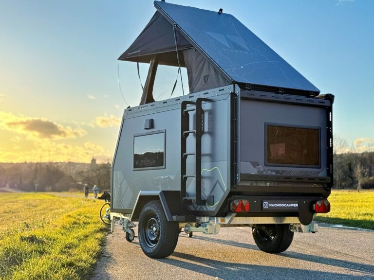 Ein kompakter Offroad-Campertrailer, das Allround-Talent von Pickupboxx, steht mit aufgebautem Aufstelldachzelt auf einer befestigten Straße am Rand eines grünen Feldes im Sonnenuntergang. Der klare Himmel taucht den maßgefertigten Pickup Camper in warmes Licht und vermittelt ein Gefühl von grenzenloser Freiheit – perfekt für Abenteurer, die einen individuell gestalteten Custom Camper suchen oder autark mit einem Pickup Camper auf Entdeckungsreise gehen möchten. Travelboxx - Pickup Wohnkabinen