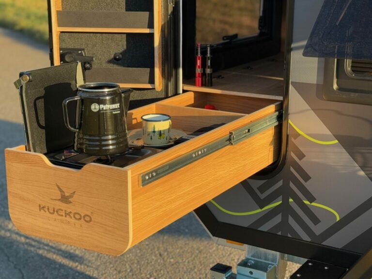 Eine ausziehbare Holzschublade eines Pickup Campers präsentiert eine stilvolle Kaffeekanne, einen Becher und einen Berg-Motiv-Becher – alles, was Sie für Ihren Outdoor-Kaffeegenuss beim Camping brauchen. Rundherum findet man durchdachte Campingausrüstung sowie einen praktischen Leiter. Der Offroad Camper trägt stolz das „Kuckoo“ Logo und moderne geometrische Designs. Die PickupBoxx ist ein echtes Allround-Talent: ideal als Custom Camper für grenzenlose Freiheit auf jedem Abenteuer abseits befestigter Straßen. Travelboxx - Pickup Wohnkabinen