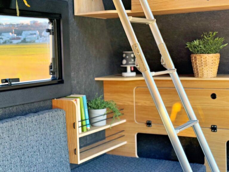 Innenraum eines Pickup Campers mit maßgefertigten Holzregalen, einer gemütlichen Sitzbank mit grauen Polstern, einem stabilen Metallleiter sowie liebevoll arrangierten Topfpflanzen und Büchern. Das Fenster eröffnet den Blick auf grenzenlose Freiheit – offene Felder und die weite Natur beladene Abenteuerlustige ein. Diese Offroad Camper Ausstattung vereint praktischen Stauraum mit individuellem Wohnkomfort und macht den Custom Camper zum idealen Allround-Talent für Reisen abseits ausgetretener Pfade. Travelboxx - Pickup Wohnkabinen