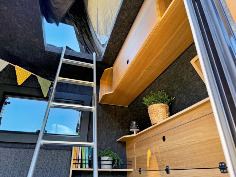 Ein gemütlicher Innenraum eines Pickup-Campers mit Holzregalen, Topfpflanzen, einer kleinen Laterne und einer Leiter, die zum Aufstelldach führt. Sonnenlicht fällt durch das Fenster und den hochgeklappten Dachbereich und unterstreicht die grenzenlose Freiheit, die dieser Allrounder von Offroad-Camper bietet. An der Wand hängt eine gelbe Wimpelkette – perfekt für alle, die einen individuellen Custom Camper suchen und Abenteuer fernab befestigter Straßen genießen möchten. Travelboxx - Pickup Wohnkabinen