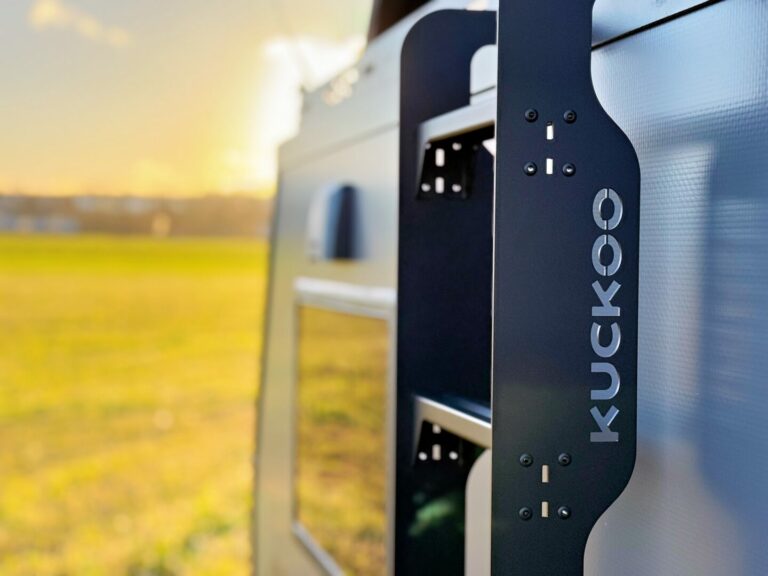Nahaufnahme eines schwarzen Metallträgers mit dem Schriftzug „KUCKOO“, montiert an der Seite eines silbernen Pickup-Campers. Dieses Bild präsentiert die Vielseitigkeit des Offroad-Campers und Custom Campers, perfekt für Abenteuer abseits befestigter Straßen. Im Hintergrund sorgt eine unscharfe, grüne Wiese im warmen Sonnenlicht für eine einladende Outdoor-Atmosphäre – ideal für alle, die einen Allround-Pickup-Camper suchen. Travelboxx - Pickup Wohnkabinen