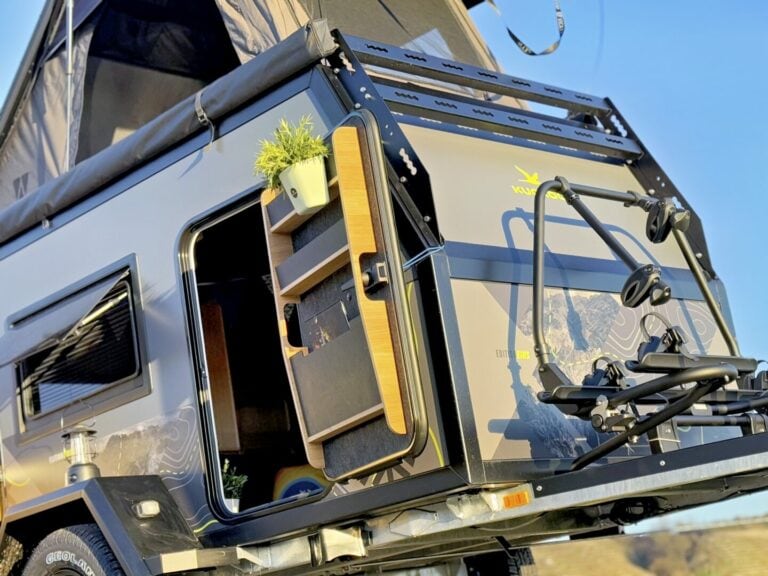 Eine Nahaufnahme eines modernen Pickup Campers mit geöffneter Seitentür zeigt stilvolle Holzverkleidungen, eine kleine Zimmerpflanze und das ausgeklappte Aufstelldach – ein echtes Offroad Camper Allround-Talent, das grenzenlose Freiheit auf jeder Reise bietet. Am Heck ist ein Fahrradträger montiert. Diese Custom Camper Lösung verbindet Komfort, Flexibilität und Abenteuerlust für alle, die unabhängig und individuell unterwegs sein möchten. Travelboxx - Pickup Wohnkabinen
