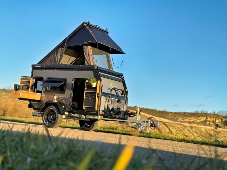 Ein stilvoller schwarzer PickupBoxx Offroad-Camper-Anhänger mit einem erhöhten Dachzelt steht auf einer malerischen Landstraße – das perfekte Symbol für grenzenlose Freiheit. Mit seiner robusten Bauweise und dem durchdachten Custom-Ausbau eignet sich dieser Pickup Camper ideal für Abenteuer abseits befestigter Wege. Umgeben von weiten Wiesen und sanften Hügeln, bietet dieser Offroad Camper alles, was Outdoor-Fans für unabhängiges Reisen unter freiem Himmel brauchen. Travelboxx - Pickup Wohnkabinen