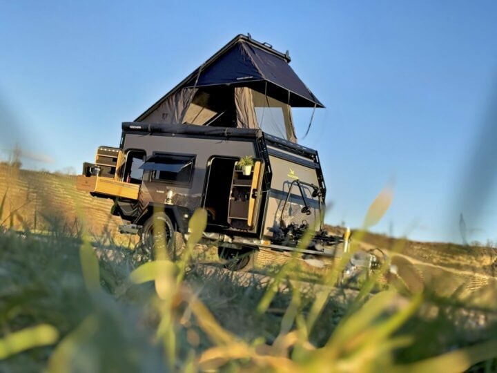 Schwarzer Offroad-Camper-Anhänger – das Allround-Talent für Abenteurer: Mit Dachzelt steht er bereit im grünen Gras. Geräumige Staufächer und eine Outdoor-Küche bieten grenzenlose Freiheit unter klarem, blauen Himmel. Ideal als Pickup Camper oder maßgeschneiderter Custom Camper für dein nächstes Offroad-Erlebnis!. Travelboxx - Pickup Wohnkabinen