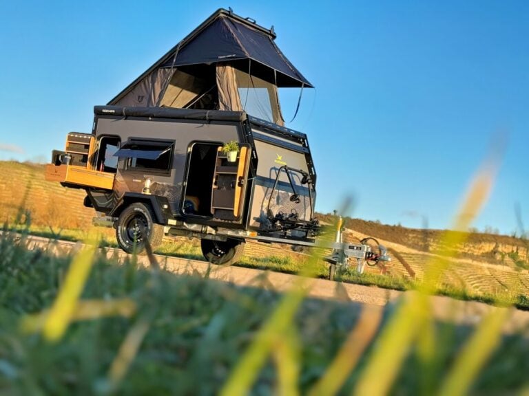 Ein schwarzer Offroad-Campertrailer – ein wahres Allroundtalent für Abenteurer – steht mit aufgestelltem Dachzelt auf einer grünen Wiese unter strahlend blauem Himmel. Die geöffneten Türen geben den Blick frei auf eine durchdachte Küche und großzügigen Stauraum, wie man es sich von einem echten Custom Camper wünscht. Im Hintergrund erstrecken sich sanfte Hügel und vermitteln das Gefühl grenzenloser Freiheit – ideal für alle, die mit einem Pickup Camper oder Offroad Camper unabhängig unterwegs sein möchten. Travelboxx - Pickup Wohnkabinen