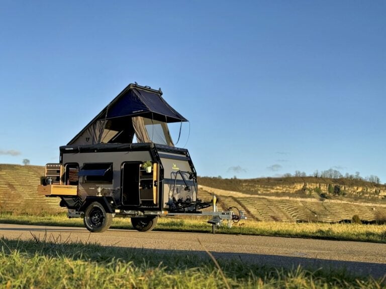 Ein schwarzer PickupBoxx Offroad-Camperanhänger mit aufstellbarem Dachzelt steht auf einer Straße – bereit für dein nächstes Abenteuer. Dieser vielseitige Pickup Camper bietet dir eine durchdachte Outdoor-Küche und jede Menge Stauraum, perfekt für grenzenlose Freiheit abseits befestigter Wege. Mit seinem robusten Design ist dieser Custom Camper die ideale Wahl für Offroad-Enthusiasten, die Komfort und Flexibilität schätzen. Eingebettet in eine Landschaft aus grünen Hügeln und strahlend blauem Himmel wird jeder Trip zum unvergesslichen Erlebnis. Travelboxx - Pickup Wohnkabinen