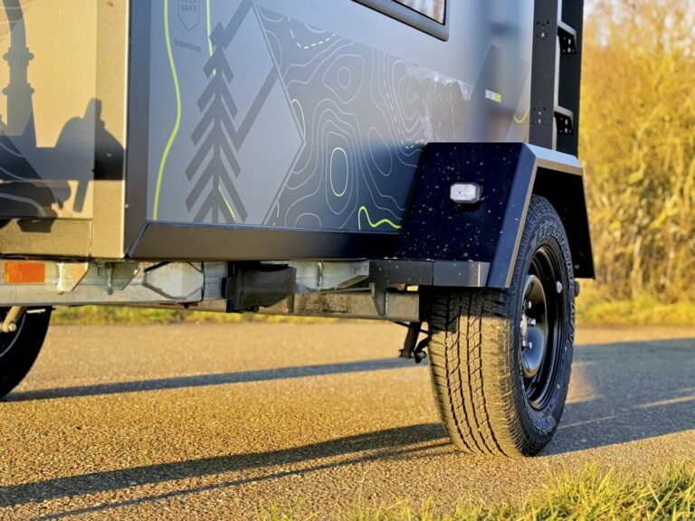Nahaufnahme eines Anhängerrads und der Seitenwand eines Pickup Campers mit modernem, karteninspiriertem Grafikdesign. Der Offroad Camper – ein echtes Allround-Talent – steht auf einer asphaltierten Straße, während Sonnenlicht darauf glänzt und im Vordergrund grünes Gras zu sehen ist. Perfekt für alle, die einen robusten Custom Camper für Abenteuer oder als vielseitigen Pickup Camper suchen. Travelboxx - Pickup Wohnkabinen