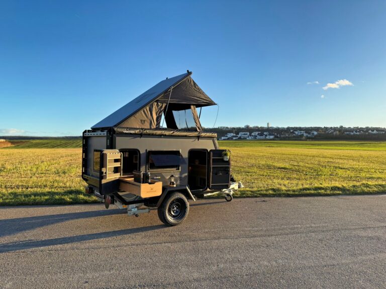 Ein kompakter, schwarzer Pickup-Camper – ein vielseitiges Offroad-Camper-Allroundtalent mit ausziehbarem Dachzelt – steht am Rand einer grünen Wiese unter strahlend blauem Himmel. Dieser maßgeschneiderte Custom Camper bietet grenzenlose Freiheit für Abenteuer abseits der Straßen, während am Horizont eine ferne Ortschaft zu sehen ist. Travelboxx - Pickup Wohnkabinen