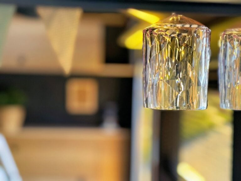 Nahaufnahme eines modernen Pickup-Campers mit strukturiertem Glas-Pendelleuchten-Design und unscharfem Hintergrund in warmen Tönen – ein wahres Allround-Talent für jedes Offroad-Abenteuer. Das sanfte, indirekte Licht schafft eine angenehme Atmosphäre im Innenraum dieses Custom-Campers. Im oberen linken Bildbereich ist leicht Wimpelgirlande zu erkennen – ideal für alle, die Stil und Komfort beim Camper-Tuning schätzen. Travelboxx - Pickup Wohnkabinen