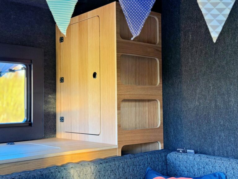 Ein kleiner, offener Holzschrank ist geschickt in die Ecke eines gemütlichen Pickup Campers integriert – ein echtes Allround-Talent für unterwegs. Über dem Offroad-Camper schwebt eine dreieckige Wimpelkette, während im Vordergrund ein Fenster und eine grau gepolsterte Sitzbank zur grenzenlosen Freiheit einladen. Perfekt für alle, die einen individuellen Custom Camper suchen und auf der nächsten Tour Komfort mit cleverem Stauraum verbinden möchten. Travelboxx - Pickup Wohnkabinen