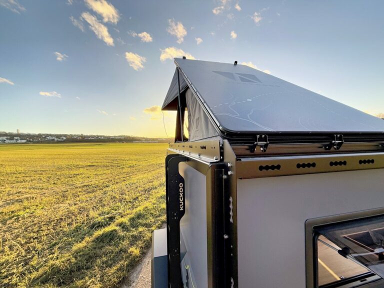 Ein modernes, aufklappbares Dachzelt thront auf einem Pickupboxx Offroad-Camper-Anhänger, der am Rand eines weiten offenen Feldes unter blauem Himmel steht. Das Sonnenlicht taucht den maßgefertigten Camper in einen warmen Schein und verspricht grenzenlose Freiheit sowie Abenteuer für alle, die mit einem Pickup Camper unterwegs sind – egal ob abseits befestigter Straßen oder beim Camping unter den Wolken. Travelboxx - Pickup Wohnkabinen