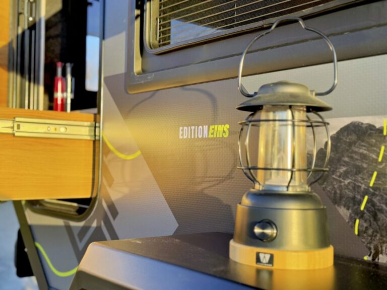 Nahaufnahme einer Laterne auf einer Ablage vor einem modernen Pickup Camper mit der Aufschrift „EDITION EVMS“ auf der Seite. Das Sonnenlicht taucht die Szene in warmen Tönen, hebt Campingausrüstung und ein geöffnetes Fenster hervor – und fängt das Gefühl grenzenloser Freiheit ein, das Offroad-Abenteuer mit einem individuell angefertigten Custom Camper bieten kann. Travelboxx - Pickup Wohnkabinen