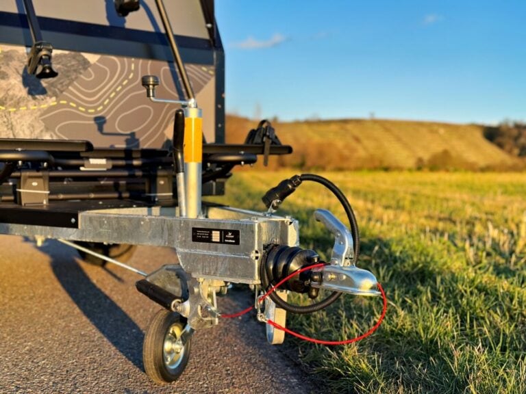 Nahaufnahme einer Anhängerkupplung und eines Rads auf einer asphaltierten Straße – im Hintergrund eine grasbewachsene Wiese und sanfte Hügel unter klarem, blauen Himmel. Der Offroad-Camper, ein Allround-Talent PickupBoxx, eignet sich ideal als Pickup Camper oder Custom Camper für alle Abenteuerlustigen. Die markante Seitenfläche des Campers ist mit einer Karte gestaltet und macht ihn zum Hingucker für Individualisten, die auf der Suche nach einem vielseitigen Camper Trailer oder einem maßgeschneiderten Pickup-Aufbau sind. Travelboxx - Pickup Wohnkabinen
