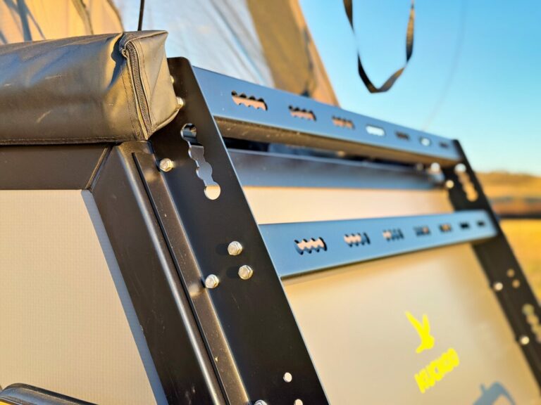 Nahaufnahme eines schwarzen Metall-Dachträgers oder Montagehalters von Pickupboxx, der an einem Fahrzeug befestigt ist. Deutlich sichtbar sind Schrauben, Schlitze und ein gepolsterter Bereich – ideal für dein Pickup Camper-Abenteuer. Der Hintergrund ist unscharf und zeigt warmes Outdoor-Licht – perfekt für grenzenlose Freiheit bei jedem Offroad Camper-Trip oder individuell ausgebauten Custom Camper. Travelboxx - Pickup Wohnkabinen