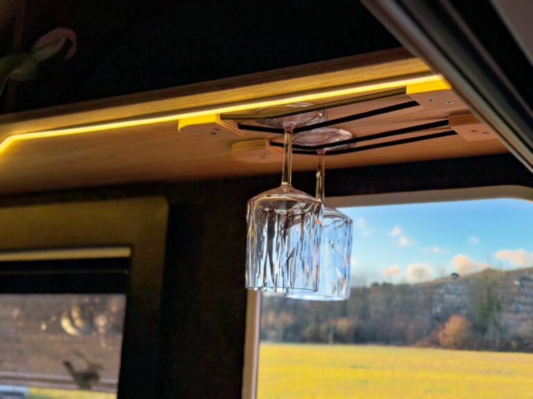 Zwei umgedrehte Weingläser hängen an einem Holzregal im Inneren eines Pickup Campers von PickupBoxx. Durch das Fenster fällt Sonnenlicht hierher und gibt den Blick auf die Natur frei – ideal, um grenzenlose Freiheit mit deinem Offroad Camper oder individuell gebauten Custom Camper unterwegs zu genießen. Travelboxx - Pickup Wohnkabinen