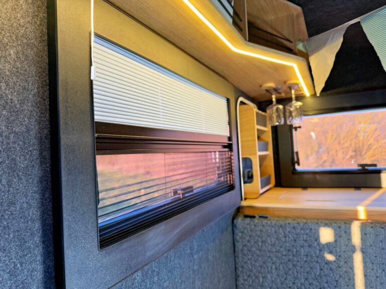 Nahaufnahme eines modernen Pickup-Camper-Innenraums: Zu sehen sind ein Fenster mit Jalousien, stimmungsvolle Unterbaubeleuchtung sowie eine stilvolle Holzarbeitsplatte mit hängenden Gläsern und dekorativer Wimpelkette. Durch das Fenster strömt hier warmes Sonnenlicht – ideal für grenzenlose Freiheit auf Offroad-Abenteuern! Egal ob individueller Custom Camper, robuster Offroad Camper oder vielseitiger Pickup Camper: So genießen Sie Komfort und Gemütlichkeit unterwegs. Travelboxx - Pickup Wohnkabinen