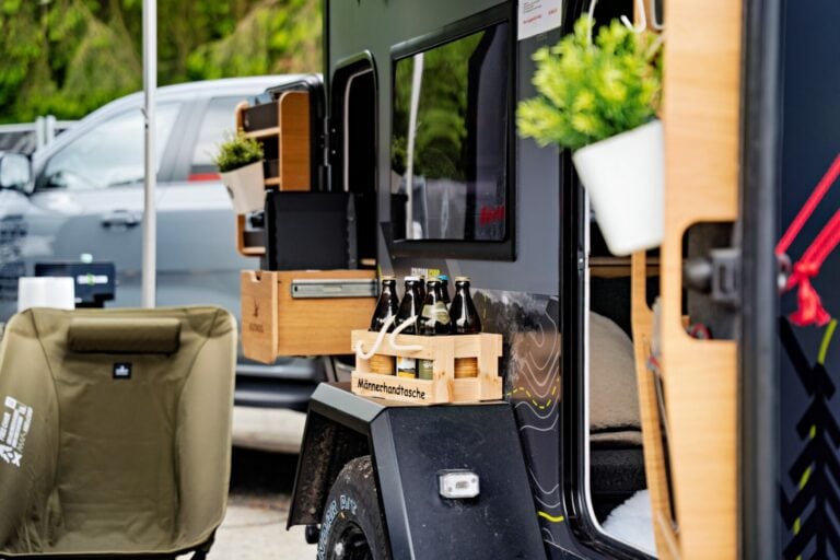 Eine Nahaufnahme eines Pickup Campers mit geöffneter Tür zeigt Regale mit Topfpflanzen und eine Holzkiste, die Bierflaschen enthält. Ein grüner Campingstuhl steht direkt davor bereit, während im Hintergrund das Zugfahrzeug geparkt ist. Die Szene vermittelt gemütliche Tiny-House-Atmosphäre – perfekt für Abenteuer im Offroad Camper oder individuell gestalteten Custom Camper. Travelboxx - Pickup Wohnkabinen