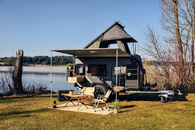 Ein moderner Travelboxx Offroad-Camper mit Aufstelldach steht idyllisch an einem See. Eine Markise verbringt Schatten über einem gemütlichen Outdoor-Essbereich mit zwei Stühlen, einem Tisch und Topfpflanzen. Umgeben von Bäumen und ruhigem Wasser wird dieser Custom Camper zur idealen Tiny House Oase für Individualisten, die einen Pickup Camper suchen und unabhängig in der Natur unterwegs sein möchten. Travelboxx - Pickup Wohnkabinen