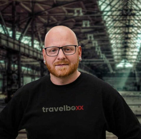 Ein glatzköpfiger Mann mit rotem Bart, Brille und einem schwarzen „travelboxx“ Sweatshirt steht vor einer großen Industriehalle mit Glasdach – perfekt für alle, die sich für Pickup Camper, Offroad Camper oder individuell angepasste Custom Camper interessieren. Travelboxx - Pickup Wohnkabinen